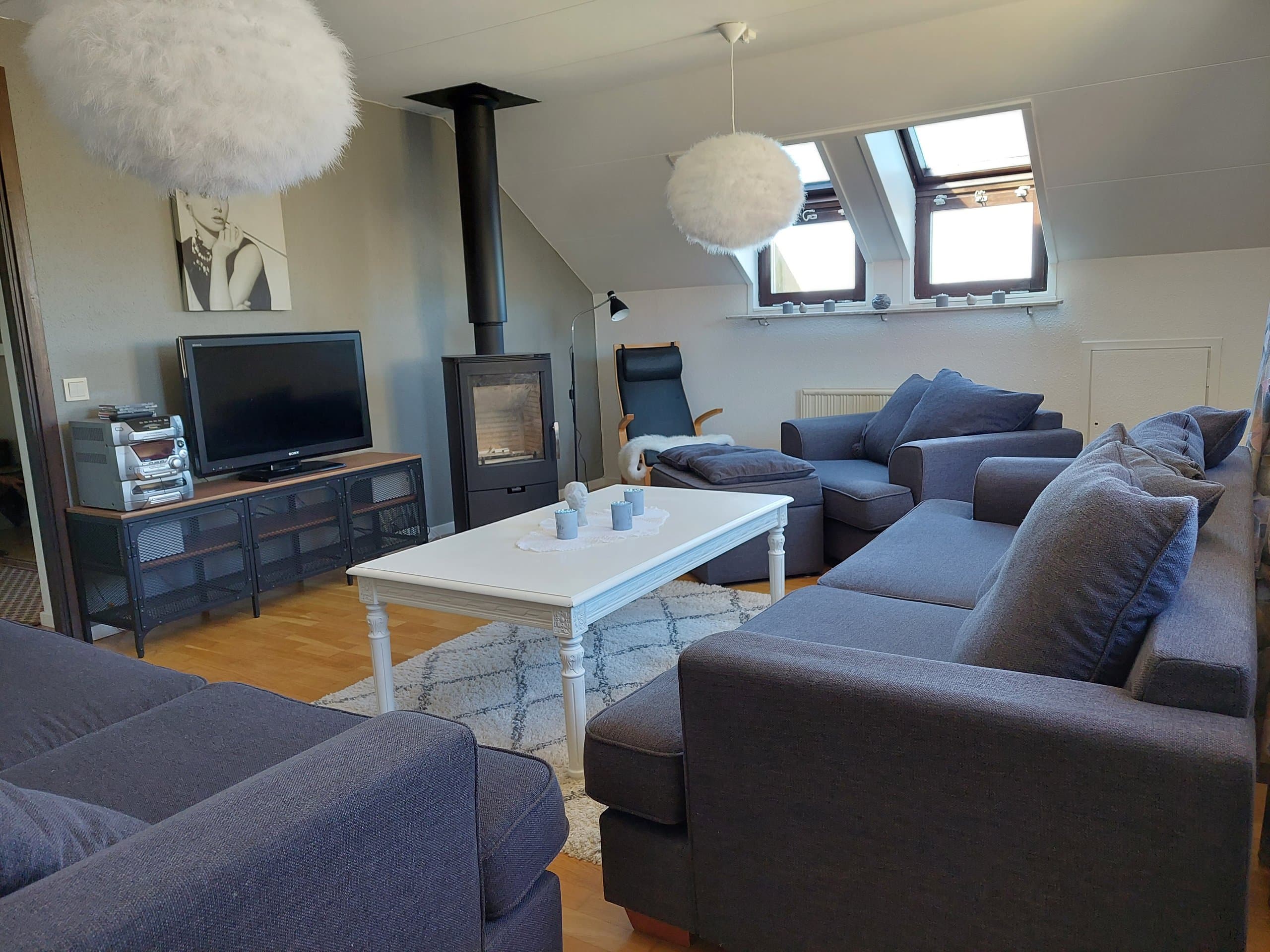 Ljust vardagsrum under snedtak med mysig vedkamin, mörkblå soffor och fåtölj, vitt soffbord och platt-TV. Takfönster släpper in dagsljus – perfekt för att samlas kring eld och TV efter en dag i spåren eller på gågatan.